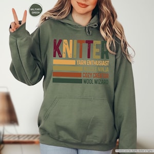 May include: A green hooded sweatshirt with the word "KNITTER" in a vintage style font. The text is layered with colorful stripes and the words "YARN ENTHUSIAST", "NEEDLE NINJA", "COZY CREATOR", and "WOOL WIZARD" are listed below.