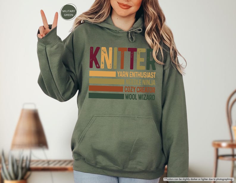 May include: A green hooded sweatshirt with the word "KNITTER" in a vintage style font. The text is layered with colorful stripes and the words "YARN ENTHUSIAST", "NEEDLE NINJA", "COZY CREATOR", and "WOOL WIZARD" are listed below.