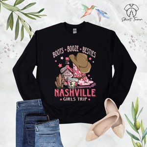 May include: Black sweatshirt with a pink and brown graphic design that says "Boots Booze Besties Nashville Girls Trip". The graphic includes a bottle of whiskey, a cactus, a glass of whiskey, and a pair of pink cowboy boots.
