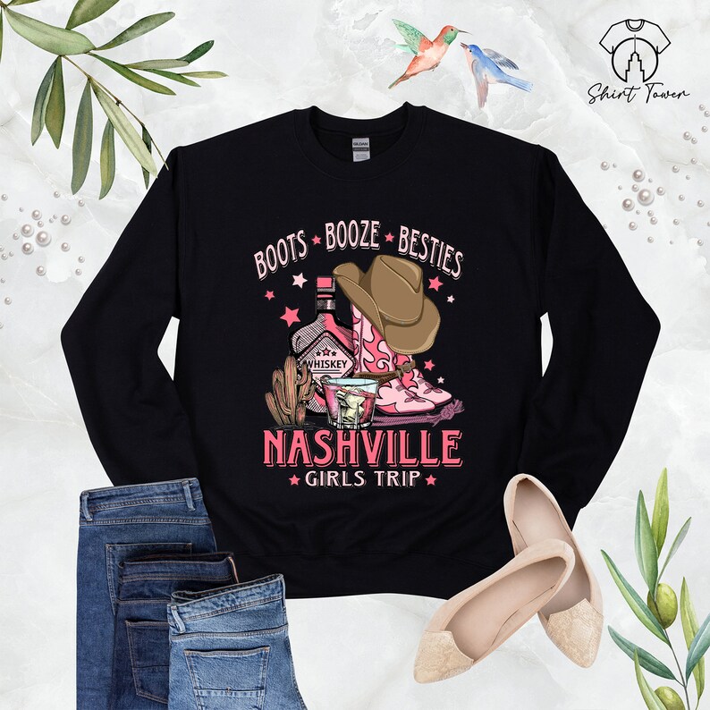 May include: Black sweatshirt with a pink and brown graphic design that says "Boots Booze Besties Nashville Girls Trip". The graphic includes a bottle of whiskey, a cactus, a glass of whiskey, and a pair of pink cowboy boots.
