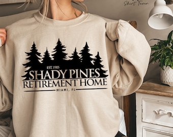 Puede incluir: Una sudadera beige con un estampado gráfico negro de una escena de bosque y el texto "Shady Pines Retirement Home - Miami, FL - Est. 1985".