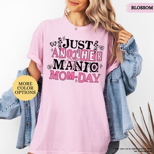May include: A light pink t-shirt with the text "JUST ANOTHER MANIC MOM-DAY" in black and pink. The shirt has a crew neck and short sleeves. The word "BLOSSOM" is in the upper right corner. A button with "MORE COLOR OPTIONS" is in the lower left.