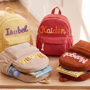 May include: Four corduroy backpacks in yellow, red, beige, and brown. Each bag features a top handle, a front zippered pocket, and a personalised name embroidered in contrasting colours: Isabel, Kaiden, Bonnie, and Margot. The bags are shown with books and a water bottle inside.