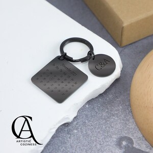 May include: Black metal keychain with a square calendar charm featuring the month of November 2023 and a heart on the 15th. The keychain also has a round charm with the initials "C&A".