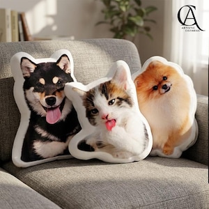 May include: Three pet-themed cushions on a grey sofa. The cushions feature realistic images of a black and tan Shiba Inu dog, a calico cat, and a Pomeranian dog. The cushions have white borders and are shaped to match the animal outlines.