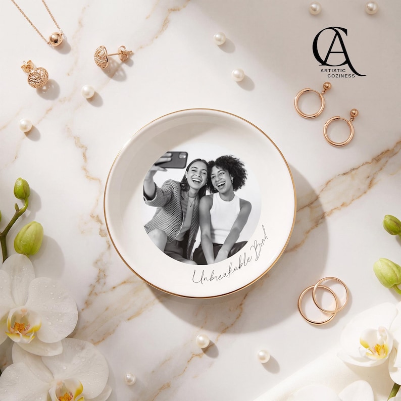 May include: A round white ceramic trinket dish with a gold-toned rim, featuring a black and white photograph of two people taking a selfie and the cursive text Unbreakable Bond printed on the surface, surrounded by scattered jewelry and pearls.