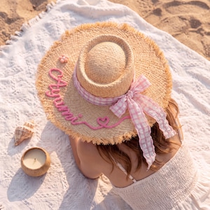 May include: A straw sun hat with a frayed edge, featuring a pink gingham ribbon tied in a bow around the crown. The hat is decorated with pink yarn lettering that reads Ayumi, a small pink flower accent, and a pink yarn heart shape.