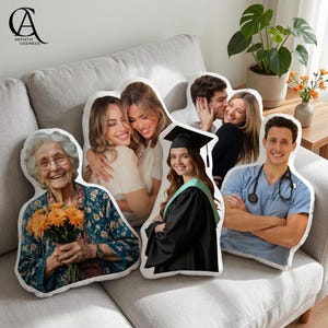 May include: A selection of custom-shaped cushions displaying photographs of individuals in diverse scenarios. The cushions showcase a woman with flowers, two women embracing, a couple, a graduate in a gown, and a man in medical attire. They are arranged on a grey sofa.