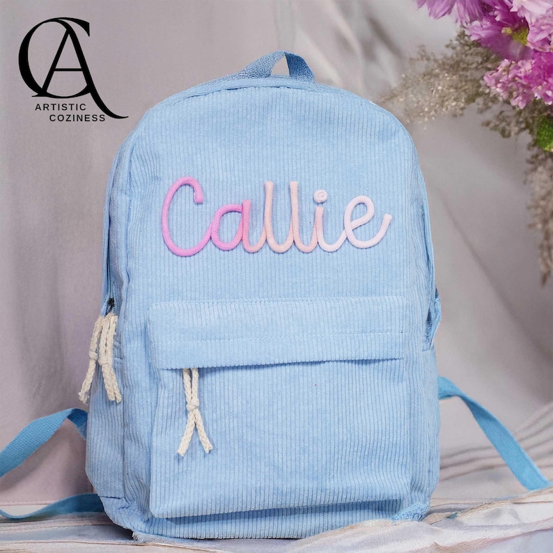 May include: A light blue corduroy backpack with a pink and white embroidered name "Callie" on the front.