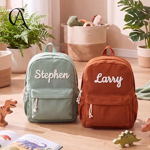 May include: Two corduroy backpacks, one sage green and one rust orange, each featuring a front zippered pocket and white embroidered names. The green backpack displays the name Stephen, while the orange backpack displays the name Larry in script.