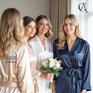 Puede incluir: Cuatro mujeres con batas de satén en tonos champán, blanco y azul marino. Las batas tienen nombres personalizados y la palabra "Bridesmaid" en la espalda. Una mujer sostiene un ramo de rosas blancas.