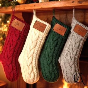 May include: Four cable-knit Christmas stockings in red, cream, green, and grey hang from a wooden mantel. Each stocking has a leather patch with a name: Adrian, Oscar, Dad, and Mom. A festive holiday decoration.
