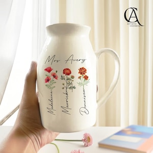 Peut inclure: Pichet en céramique crème avec une anse incurvée, orné d'illustrations florales et des noms "Madeleine", "Mavrick" et "Dennizsa" sous les mots "Mrs. Avery". Le pichet a une large ouverture et est tenu par une main.