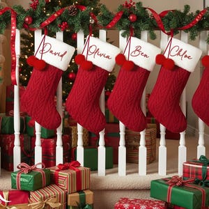 May include: Five red Christmas stockings with white cuffs and names embroidered on them hang from a garland-decorated railing. Each stocking has a red pom-pom. Presents wrapped in green, red, and gold are stacked below.