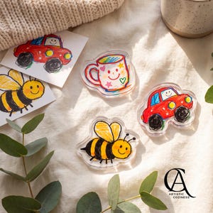 May include: Clear acrylic charms featuring colourful children's drawings of a red car, a yellow and black bumblebee, and a white mug with a smiley face. The set includes matching paper prints of the car and bee designs, all arranged together.
