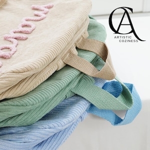 May include: A stack of four fabric bags in different colors, including beige, green, and blue. The beige bag has the word "good" embroidered on it in pink thread. The bags have fabric handles.