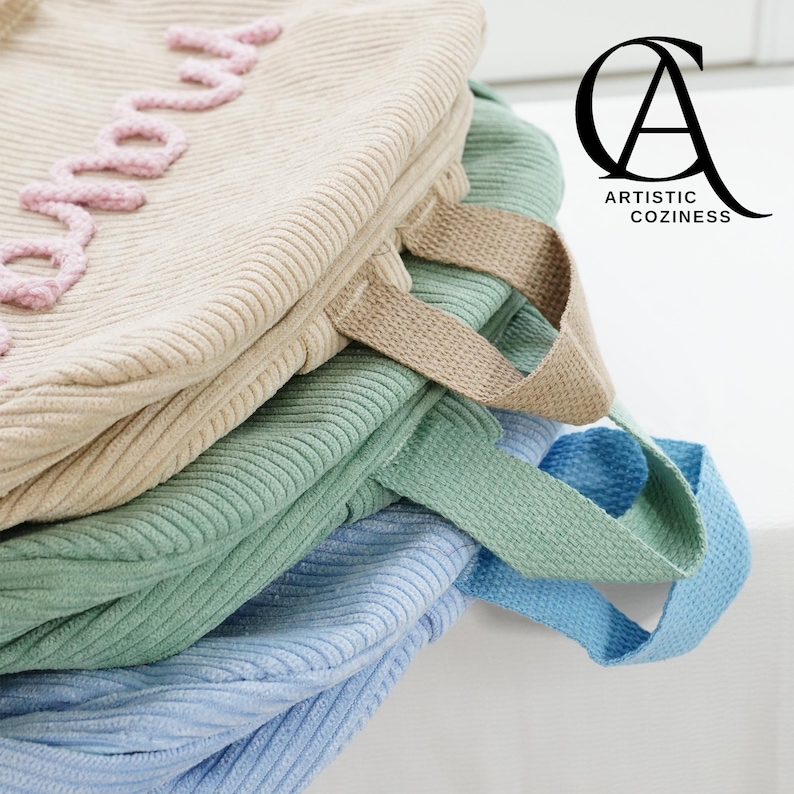 May include: A stack of four fabric bags in different colors, including beige, green, and blue. The beige bag has the word "good" embroidered on it in pink thread. The bags have fabric handles.