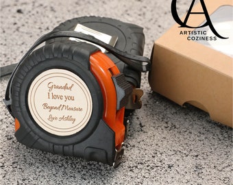 Puede incluir: Una cinta métrica negra y naranja con un círculo de madera unido al frente. El círculo tiene el texto "Grandad I love you Beyond Measure Love Ashley" grabado.