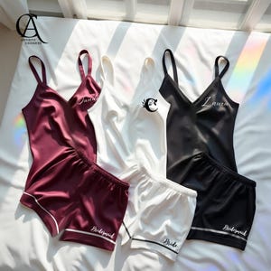 Könnte beinhalten: Satin-Schlafanzüge in Burgunderrot, Weiß und Schwarz. Jedes Set enthält ein Camisole und Shorts. Das burgunderfarbene Set hat "Jane" auf dem Camisole und "Bridesmaid" auf den Shorts. Das weiße Set hat "Bride" auf den Shorts. Das schwarze Set hat "Laura" auf dem Camisole und "Bridesmaid" auf den Shorts.