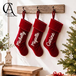 May include: Three deep red velvet Christmas stockings hanging from a rustic wooden rack with black metal hooks. Each stocking features white embroidered script lettering displaying the names "Michelly," "Miyselle," and "Carine" on the front.