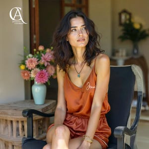 May include: Rust-colored satin pajama set with a camisole top and shorts. The top is personalized with the name "Jenni" in white script. The model is wearing a necklace and bracelets. The image includes a vase of flowers and the text "Artistic Coziness."