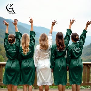 May include: Five women wearing satin robes with their arms raised. Four robes are emerald green with white text that reads "Larissa", "Chloe", "Julie", and "M". The fifth robe is white with the name "Camilla". The robes have lace trim.