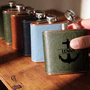 May include: A selection of leather-covered hip flasks in shades of green, brown, black, and blue. The front flask is olive green with a black anchor design and the name "LENNY". Each flask has a silver cap.