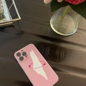 Peut inclure: Coque de téléphone rose avec un motif blanc représentant le contour d'un pays, entouré d'une orbite rose parsemée de cœurs. Le boîtier comporte un module caméra avec plusieurs lentilles. En arrière-plan, une surface sombre et un vase avec des fleurs.