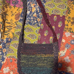 May include: A handmade, crocheted shoulder bag in a rectangular shape with a rounded bottom. The bag features a gradient of colors, including teal, green, yellow, and purple. The strap is a dark gray color with a burgundy accent. The bag is displayed on a colorful patchwork quilt.