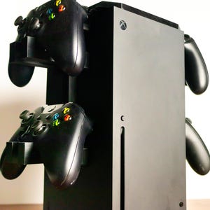 Op de afbeelding: Zwarte Xbox Series X console- en controllerstandaard. De standaard biedt ruimte voor de console en maximaal vier controllers. De controllers zijn zwart met veelkleurige knoppen. De standaard is zwart en heeft een strak, modern ontwerp.