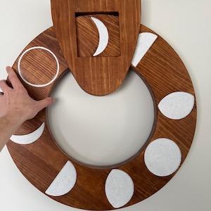May include: A wooden moon phase wall hanging. The circular design features a dark brown stained wood with white painted moon phases. The phases include crescent, half, and full moons. The top section has a square cutout with a crescent moon.