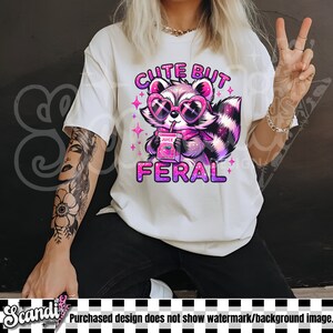 Op de afbeelding: Wit T-shirt met een roze en paarse cartoon-wasbeer die een zonnebril draagt en een sapdoos vasthoudt. De tekst "Cute But Feral" staat in roze letters boven de wasbeer.