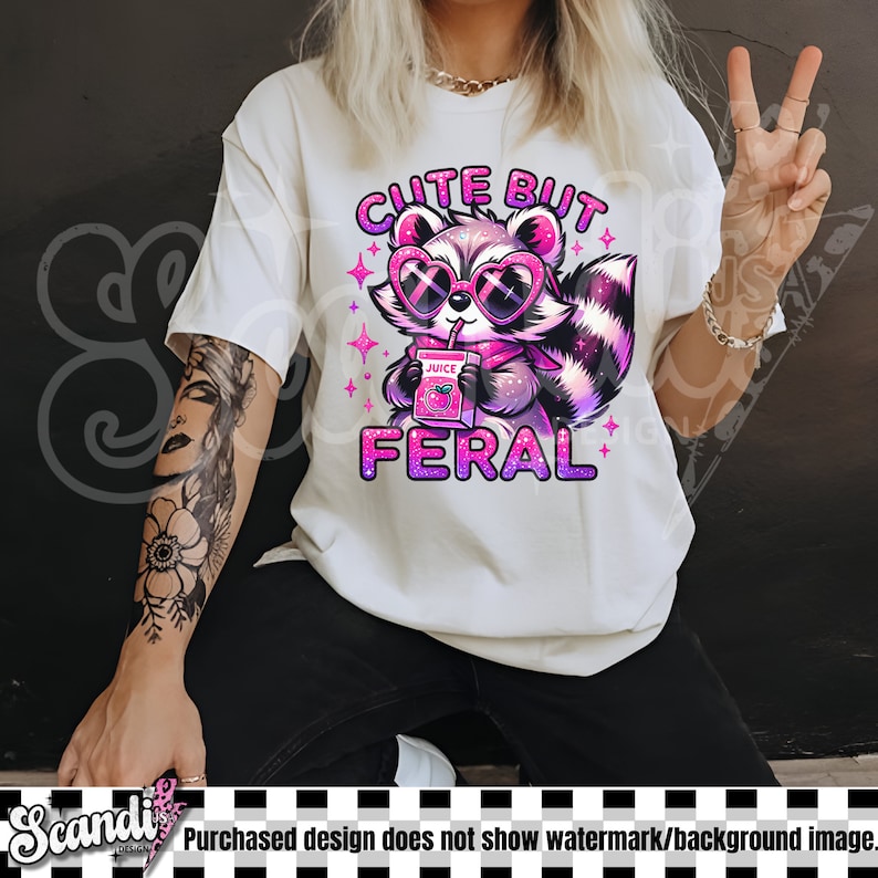 Op de afbeelding: Wit T-shirt met een roze en paarse cartoon-wasbeer die een zonnebril draagt en een sapdoos vasthoudt. De tekst "Cute But Feral" staat in roze letters boven de wasbeer.