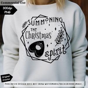 May include: A white sweatshirt with black text that reads "Summoning the Christmas spirit" in a geometric frame. The design includes a stylized Ouija board planchette and a sprig of greenery.