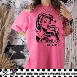 May include: A bright pink t-shirt featuring a black and white graphic of a woman drinking tea, with the text "JUST HERE FOR THE tea". The shirt has short sleeves and a relaxed fit.