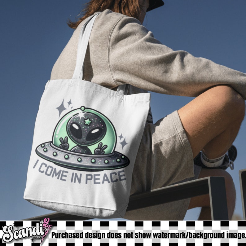 May include: A white canvas tote bag featuring a graphic of a black alien with a starry head inside a green-tinted UFO. The alien is making peace signs with both hands. Below the UFO, the text reads I COME IN PEACE in bold, dark grey letters.