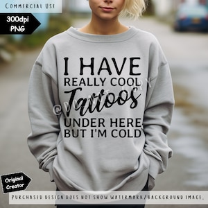 May include: A light grey crewneck sweatshirt featuring black text that reads I HAVE REALLY COOL Tattoos UNDER HERE BUT I'M COLD. The garment has long sleeves and a ribbed neckline, cuffs, and hem, providing a standard fit for casual wear.
