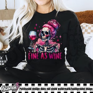 K&ouml;nnte beinhalten: Ein schwarzes Sweatshirt mit einem pinkfarbenen und wei&szlig;en Skelett-Motiv, das ein Glas Wein h&auml;lt. Der Text "FINE AS WINE" ist unter dem Motiv aufgedruckt.