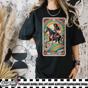 May include: Black t-shirt featuring a tarot card design with a skeleton cowgirl riding a horse over a rainbow. The card is framed with floral accents and the text "THE COWGIRL".