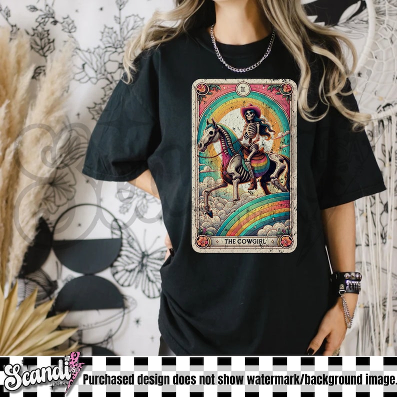 May include: Black t-shirt featuring a tarot card design with a skeleton cowgirl riding a horse over a rainbow. The card is framed with floral accents and the text "THE COWGIRL".