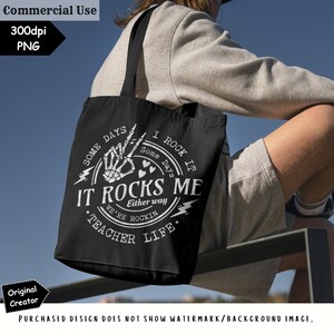 K&ouml;nnte beinhalten: Schwarze Tragetasche mit wei&szlig;em Text und Grafiken. Das Design enth&auml;lt die Worte "IT ROCKS ME" mit einer Skeletthand, die eine Rock-and-Roll-Geste macht. Der Text "TEACHER LIFE" ist ebenfalls enthalten. Die Tasche hat schwarze Tr&auml;ger.