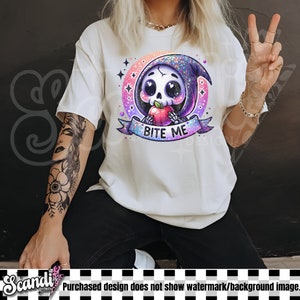 以下が含まれることがあります： クリーム色のTシャツ。赤いリンゴを持った漫画の骸骨が描かれており、「BITE ME」の文字がバナーに書かれています。デザインはカラフルでキラキラ光る三日月と星の中に配置されています。カジュアルウェアです。