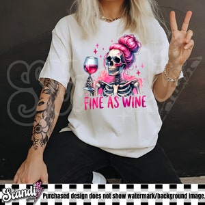 K&ouml;nnte beinhalten: Wei&szlig;es T-Shirt mit einem rosafarbenen Skelett, das ein Glas Rotwein h&auml;lt. Das Skelett hat rosafarbenes Haar in einem Dutt und der Text "Fine as Wine" ist unter dem Bild gedruckt.