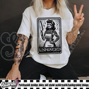 以下が含まれることがあります： ナイフを持った女性の白黒のイラストが描かれた白いTシャツ。イラストはタロットカードのスタイルで、下部に「The Unhinged」と書いてあります。