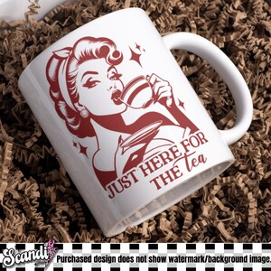 May include: White ceramic mug with a red graphic of a woman drinking tea, with the text "JUST HERE FOR THE TEA". The mug has a curved handle and is set against a brown background.