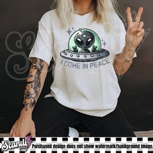 May include: A white short-sleeve t-shirt featuring a graphic of a grey alien inside a green-tinted glass dome on a silver flying saucer. The alien is making a peace sign with both hands. Below the saucer, the text reads I COME IN PEACE.