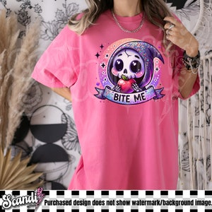 以下が含まれることがあります： ピンク色のTシャツ。漫画の死神のキャラクターが描かれています。死神は骸骨の顔、紫色のフード、赤いリンゴを持っています。「BITE ME」の文字がバナーに書かれています。デザインは星の付いた円形のフレームに入っています。
