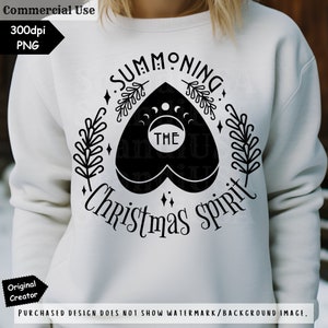 Op de afbeelding: Een wit sweatshirt met een zwart grafisch ontwerp met de tekst "Summoning The Christmas Spirit". Het ontwerp bevat een zwart hart met een wassende maan en het woord "THE" erin.