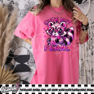 Op de afbeelding: Roze T-shirt met een afbeelding van een cartoonwasbeer met een zonnebril en een doosje sap. De tekst "Cute But Feral" is op het T-shirt gedrukt.