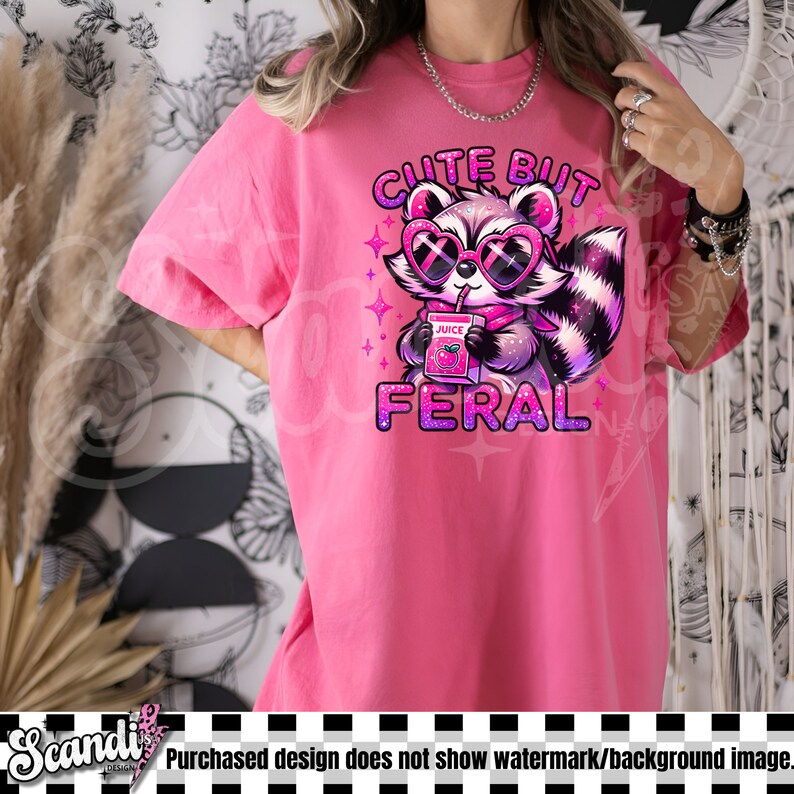 Op de afbeelding: Roze T-shirt met een afbeelding van een cartoonwasbeer met een zonnebril en een doosje sap. De tekst "Cute But Feral" is op het T-shirt gedrukt.